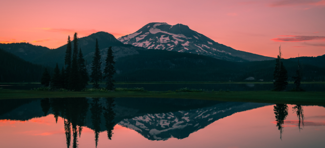 A pink sunset sky with two mountain peaks whose reflections can be seen in the still water of the lake beneath them.