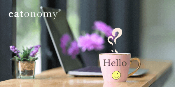 An open laptop on a desk next to a small vase of purple flowers and a pink coffee mug that says "hello" on it with a smiley face.